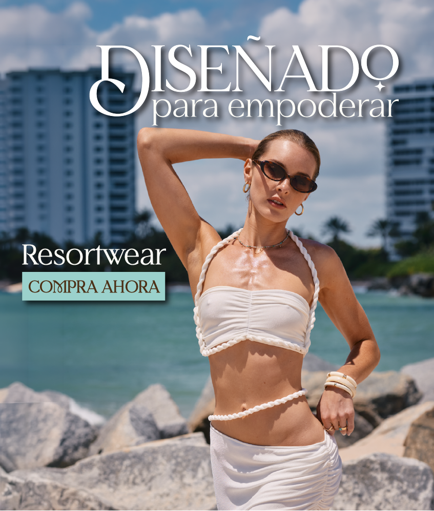 Mujer vestida de blanco y vestida de resort parada en una playa rocosa con edificios al fondo